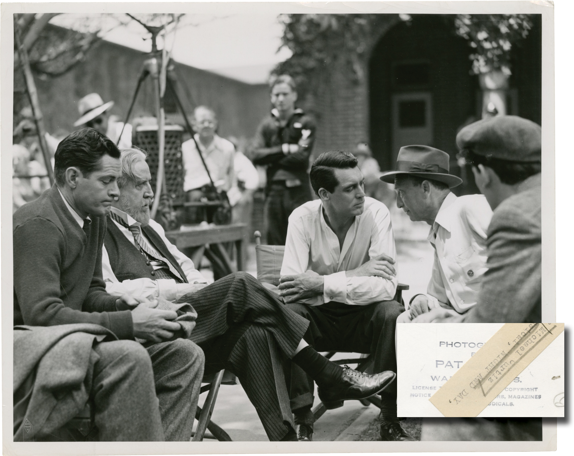 Black and white vintage photograph from the 1946 film, Night and Day, showing director Michael Curtiz, and stars Cary Grant, Monty Woolley, and Donald Woods conferring on the set. With the stamp of photographer Pat Clark and a mimeo snipe on the verso. 