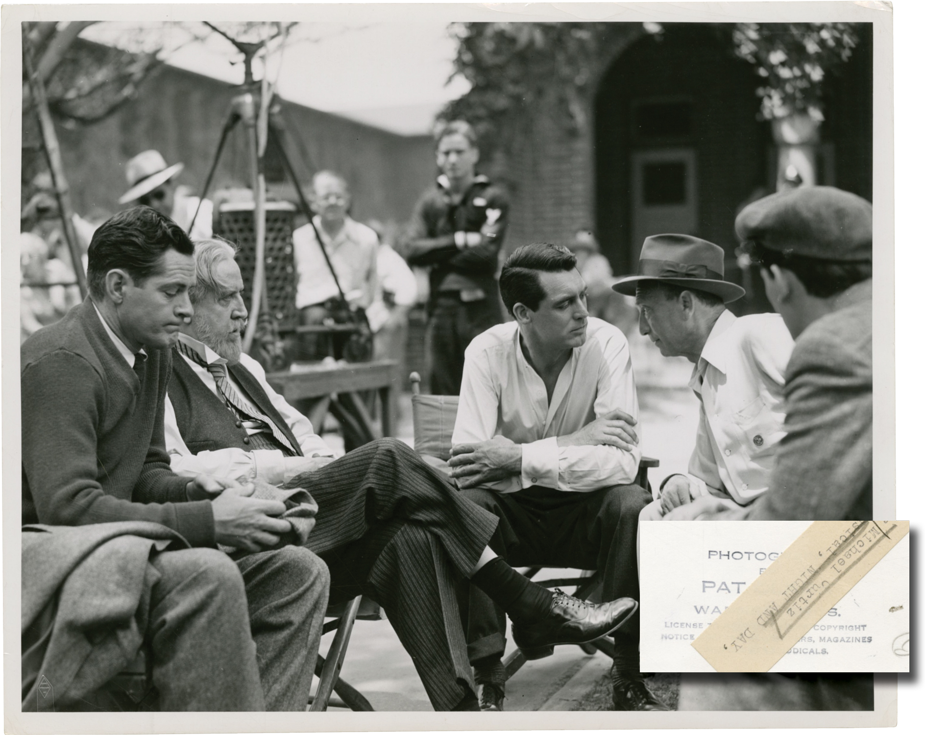 Black and white vintage photograph from the 1946 film, Night and Day, showing director Michael Curtiz, and stars Cary Grant, Monty Woolley, and Donald Woods conferring on the set. With the stamp of photographer Pat Clark and a mimeo snipe on the verso.