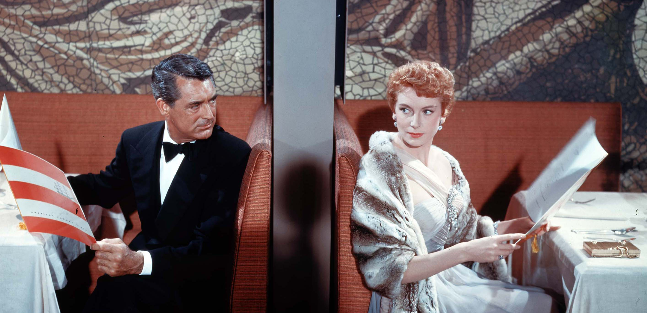 Cary Grant (Nicky Ferrante) looks quizzically at Deborah Kerr (Terry McKay) as they sit back to back in the orange booths of the trans-Atlantic ocean liner's dining room. 